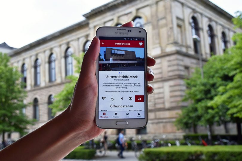 Zu sehen ist ein Handy mit der Startseite der App, das vor das Altgebäude der TU Braunschweig gehalten wird.