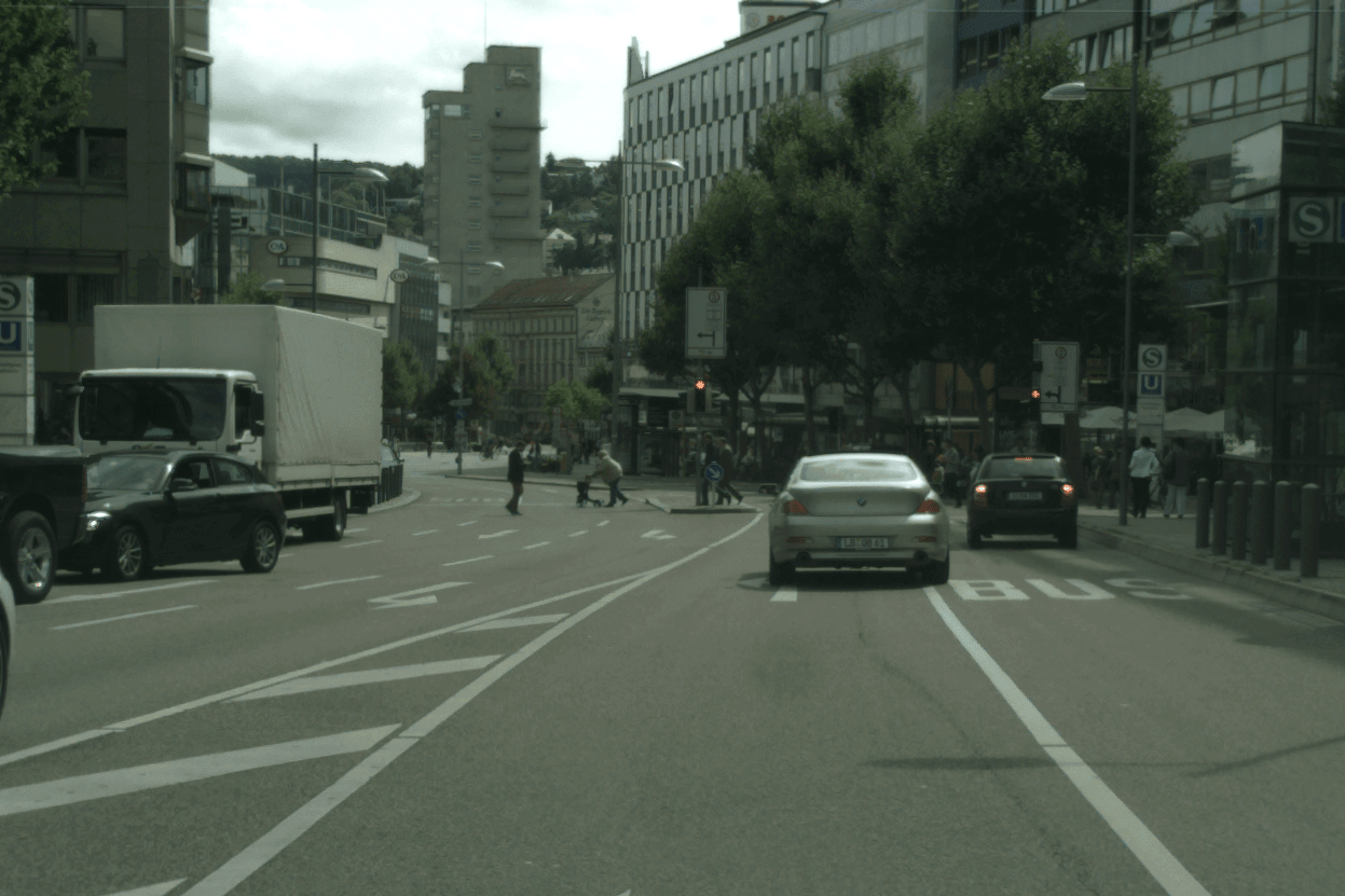Der Blick aus Perspektive des Fahrendens auf eine mehrspurige Straße. Zu sehen sind mehrere parkende und fahrende Autos, sowie eine rote Ampel, an der Fußgänger*innen kreuzen.