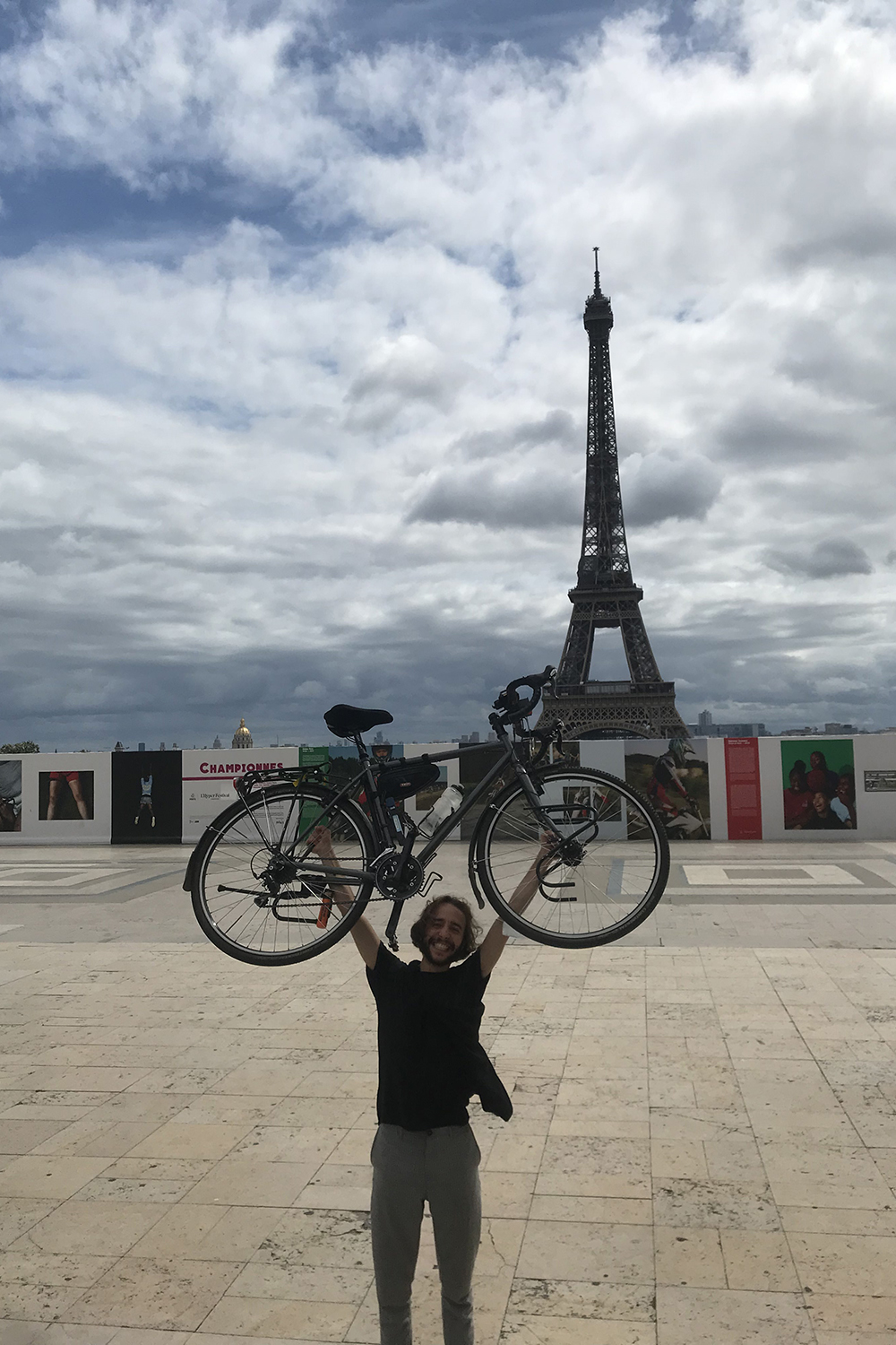 Moses Köhler holds his bicycle in the air. Behind him you can see the Eiffel Tower.