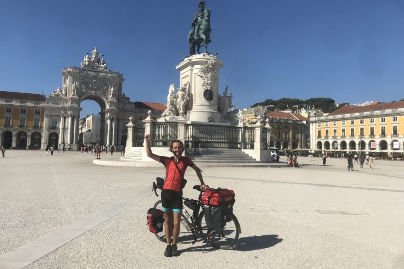 Moses Köhler stands with his bicycle in front of a landmark of the city of Lisbon.