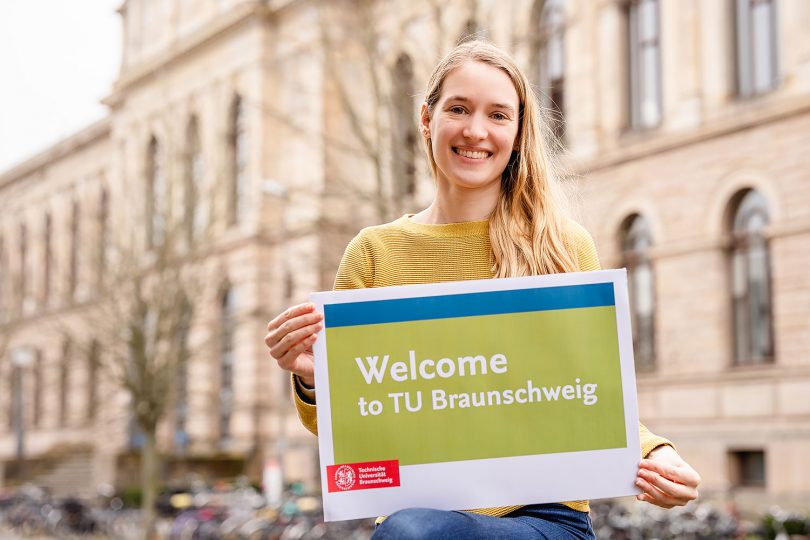 Marilena Zitka, Koordinatorin International Student Support, mit einem Schild mit dem Aufdruck „Welcome to TU Braunschweig“ vor dem Altgebäude.