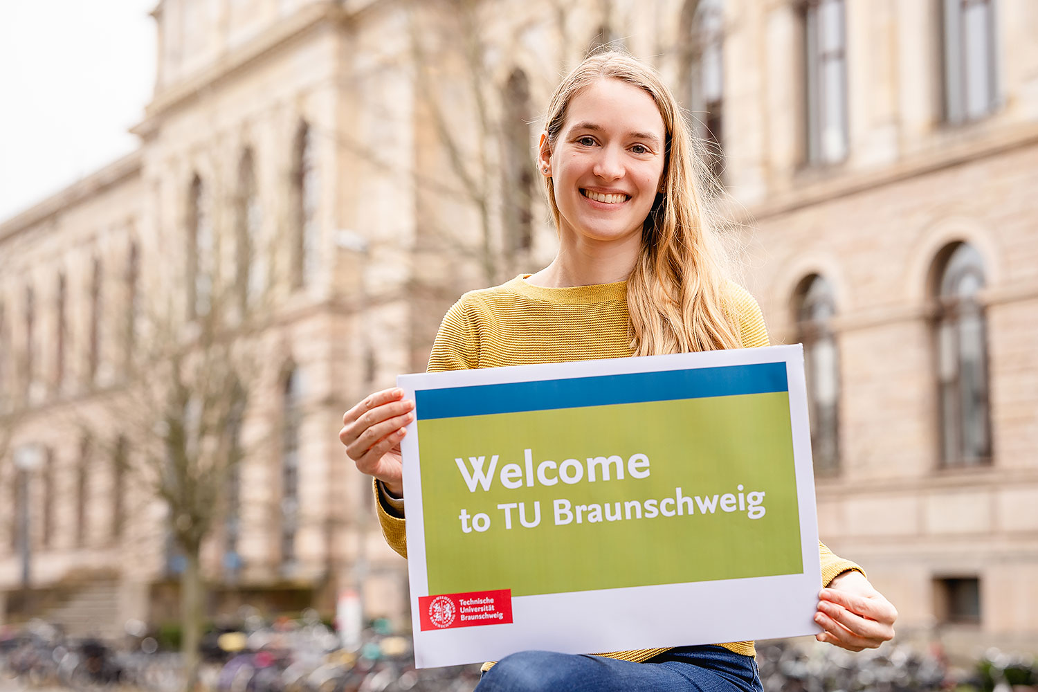 Marilena Zitka, Koordinatorin International Student Support, mit einem Schild mit dem Aufdruck „Welcome to TU Braunschweig“ vor dem Altgebäude.